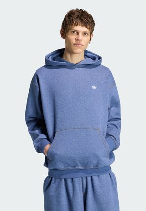 Ung mand med kort krøllet hår iført en blå Adidas hættetrøje og matchende joggingbukser, hænderne i forlommen, neutral baggrund.