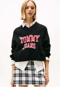 Schwarzer cropped Pullover mit "TOMMY JEANS" in pinker Textur, V-Ausschnitt, kombiniert mit einem weißen Hemd mit Kragen und einem schwarz-weißen karierten Rock.