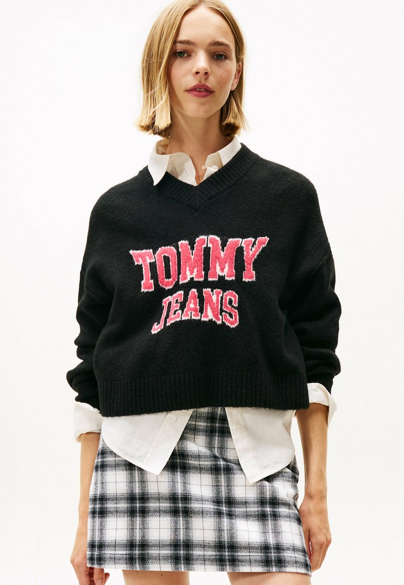 Schwarzer cropped Pullover mit "TOMMY JEANS" in pinker Textur, V-Ausschnitt, kombiniert mit einem weißen Hemd mit Kragen und einem schwarz-weißen karierten Rock.