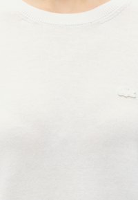 Gros plan d'une personne portant un pull en maille blanc uni avec un petit logo blanc en relief sur la poitrine.