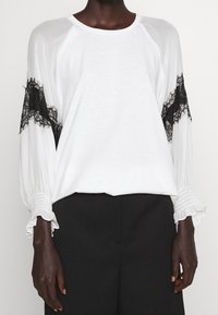 Top blanco de manga larga con detalles en encaje negro en las mangas, puños texturizados y un corte holgado. La tela parece suave y lisa.