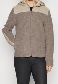 Giacca marrone in misto lana con sezione superiore e cappuccio in nylon beige. Presenta una zip frontale e tasche laterali con texture liscia.