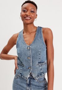 Chaleco de denim con escote en V, seis botones, dos bolsillos frontales y un diseño corto. Presenta un color azul claro con texturas desgastadas.