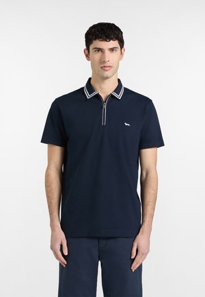 Giovane uomo frontale che indossa una polo a maniche corte blu navy con collo a zip e piccolo logo bianco di cane sul petto.