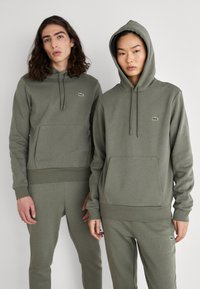 Sudaderas con capucha en verde oliva, con bolsillos frontales y un pequeño logo, combinadas con pantalones de chándal a juego. Material suave de mezcla de algodón.