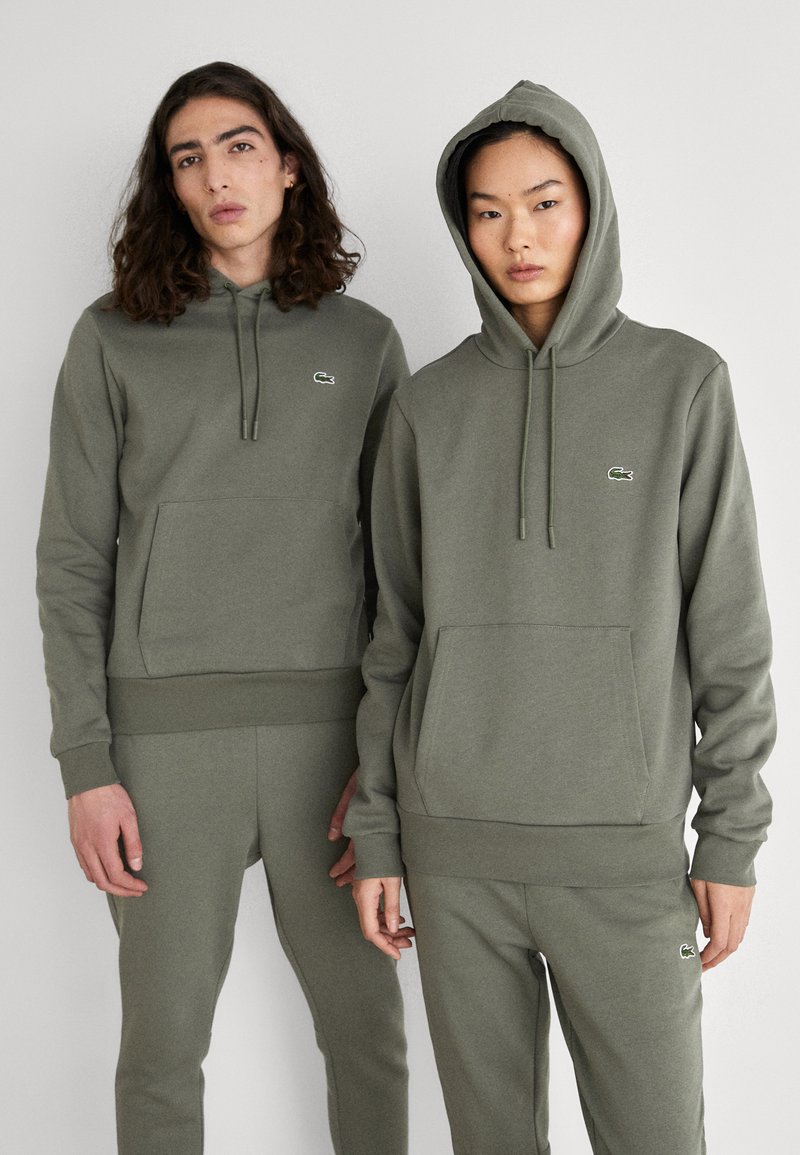Sudaderas con capucha en verde oliva, con bolsillos frontales y un pequeño logo, combinadas con pantalones de chándal a juego. Material suave de mezcla de algodón.