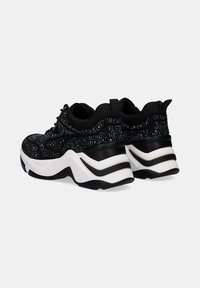 Zapatillas negras con una parte superior texturizada, que presentan acentos que brillan y una suela blanca gruesa con líneas negras. El diseño incluye una puntera redonda y un sistema de cordones en la parte delantera.