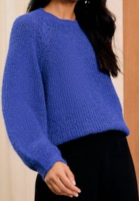 Pull bleu tricoté avec une finition texturée, col rond et manches longues légèrement bouffantes. Associé à un pantalon noir.