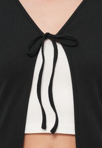 Μαύρο ριμπ ζακέτα με V-λαιμόκοψη και δέσιμο μπροστά. Διαθέτει υφή με ανάγλυφα σχέδια, ελαφρώς εφαρμοστή γραμμή και αποκαλύπτει λευκό τοπ από κάτω.