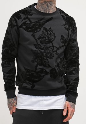 Mann trägt schwarzen Pullover mit erhabenem Samt-Floralmuster und Vogelmotiv, darunter ein weißes Hemd, Hände tätowiert und entspannt an den Seiten.