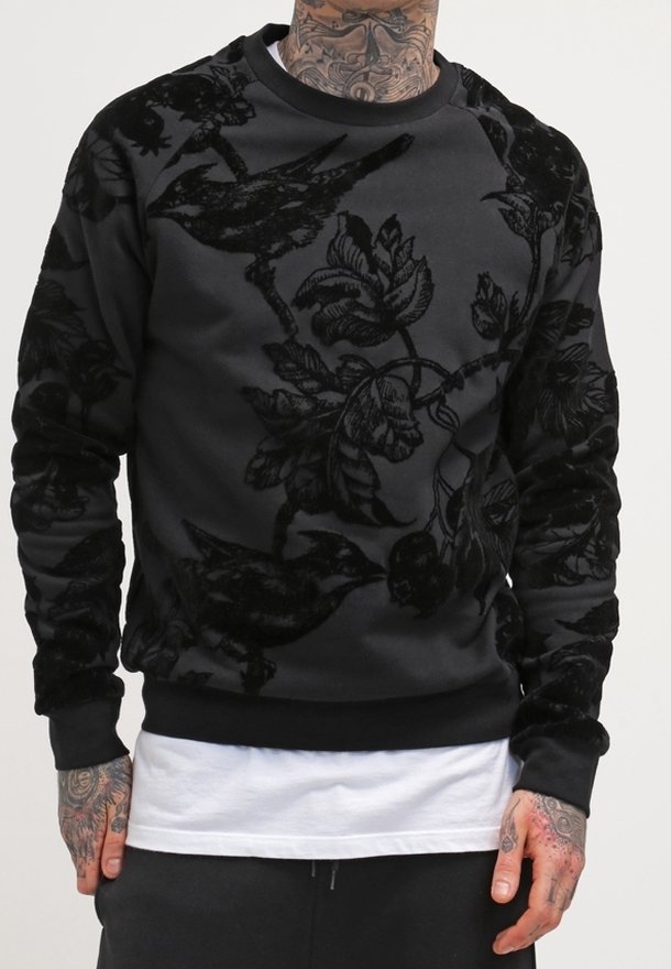 Homme portant un sweat-shirt noir orné d'un motif floral et d'oiseaux en velours surélevé, superposé à une chemise blanche, les mains tatouées et détendues le long du corps.