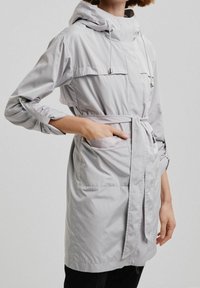 Veste gris clair mi-longue avec capuche, cordon de serrage et poignets élastiques. Dispose de poches et d'une ceinture nouée pour un ajustement réglable. Texture lisse.
