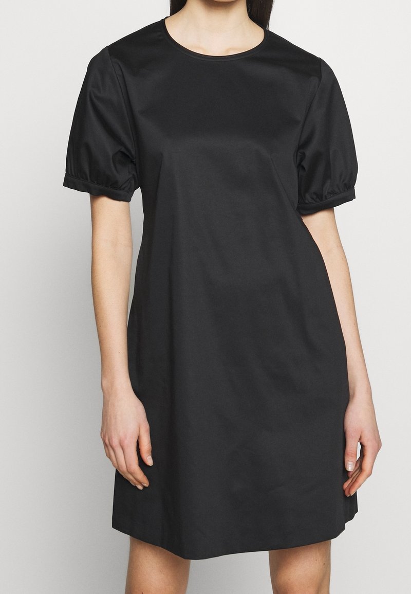 Femme portant une robe noire à manches courtes bouffantes et un col rond sur un fond clair uni.