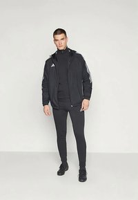 Jachetă sport neagră cu dungi albe, purtată cu un top negru ajustat și colanți. Dispune de un fermoar și accente cu logo, asortată cu pantofi negri.