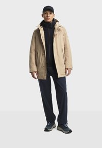 Beige waterdichte jas met een rits, zwarte binnenlaag, marineblauwe broek en grijze sneakers met blauwe accenten, gedragen door een staand persoon.