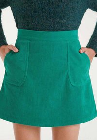 Falda A-line de pana en color verde azulado con grandes bolsillos delanteros, combinada con un suéter oscuro texturizado, con las manos metidas en los bolsillos.