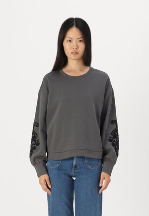 Sweatshirt gris à manches longues avec des motifs décoratifs noirs sur les manches. Texture douce et coupe décontractée avec un col rond.