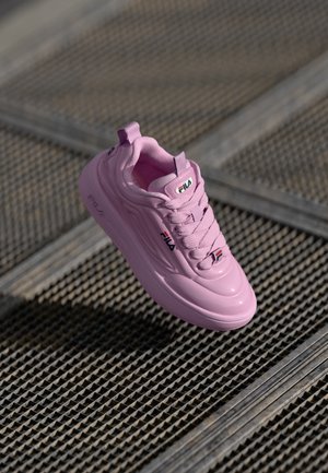 Pink athletic shoe with a glossy finish, featuring a padded tongue, lace-up front, and Fila logo. Rounded toe and structured sole design.