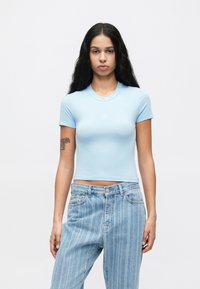 Νεαρή γυναίκα με μακριά μαύρα μαλλιά, φορώντας ένα ανοιχτό μπλε κοντομάνικο μπλουζάκι και ψηλόμεσα ριγέ τζιν, με φόντο έναν απλό τοίχο.