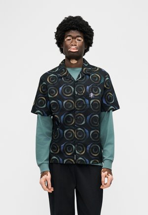 Άτομο με λεύκη που φοράει μαύρο πουκάμισο με κοντά μανίκια και κουμπιά, διακοσμημένο με κυκλικά σχέδια πάνω από πράσινο πουκάμισο με μακριά μανίκια, στέκεται μπροστά σε απλό φόντο.