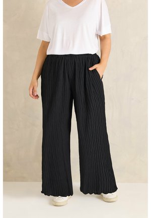 Persona usando pantalones negros anchos y texturizados, sueltos, camiseta blanca de manga corta con cuello en V y zapatillas blancas, de pie contra un fondo neutro.