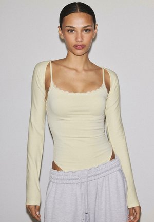 Donna con capelli tirati indietro che indossa una maglia a strati di colore giallo chiaro con spalline sottili e maniche lunghe, abbinata a pantaloni della tuta grigio chiaro.