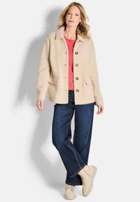 Beige, gestructureerde cardigan met knopen aan de voorkant en twee voorzakken, gedragen over een koraalkleurig topje, gecombineerd met donkerblauwe wijde jeans en beige sneakers.