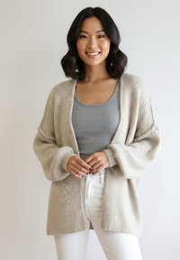 Beżowy dzianinowy kardigan o luźnym kroju, z fakturowanej tkaniny, z otwartym przodem i długimi rękawami, noszony na szarym topie i białych spodniach.