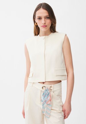 Ung kvinde iført ærmeløs cremefarvet vest og matchende højtaljede bukser med stribet tørklæde bundet ved bæltestropperne, stående mod en hvid baggrund.