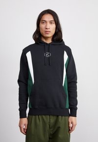 Mustapaitapaita, jossa on valkoisia ja vihreitä paneeleita, etutasku ja narusysteemi huppua varten. Materiaali näyttää pehmeältä ja rento istuvuus.