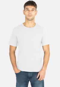 Weißes kurzärmliges T-Shirt mit Rundhalsausschnitt, aus weichem Stoff, mit dezentem Logo auf der Brust, kombiniert mit blauen Jeans.