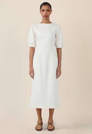 Frau trägt ein weißes Midikleid mit halblangen Puffärmeln und braunen flachen Sandalen, stehend vor einem beigen Hintergrund.