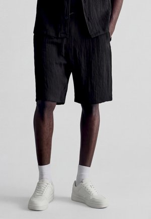 Mężczyzna w czarnych teksturowanych szortach, białych skarpetkach i białych trampkach stoi na tle jednolitego jasnego tła.