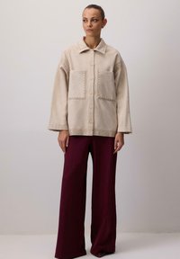 Beżowa kurtka polarowa z kołnierzem, dużymi kieszeniami i ozdobnym przeszyciem; noszona na bordo spodniami o szerokich nogawkach.