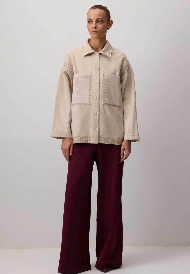 Beżowa kurtka polarowa z kołnierzem, dużymi kieszeniami i ozdobnym przeszyciem; noszona na bordo spodniami o szerokich nogawkach.