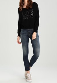 Černý oversized svetr s nápisem "LOVE" z flitrů, spárovaný s tmavě modrými skinny jeans a světlobéžovými teniskami.