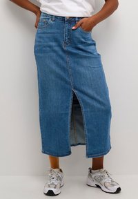 Falda vaquera azul con abertura frontal, dos bolsillos laterales y dobladillos cosidos. Combinada con zapatillas grises con detalles de malla y goma.