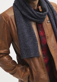 Brun læderjakke, mørkeblå ribstrikket halstørklæde, rødternet skjorte under, med en metallisk knapdetalje på jakken. Mørke, strukturerede lag.