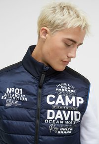 Marineblaue Steppweste mit hohem Kragen und Logo-Drucken. Der Stoff ist quilted mit sichtbaren Nähten, verfügt über einen Reißverschluss und seitliche Akzente.