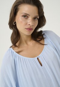 Blouse plissée bleu clair avec un col rond, des détails de bouton et des manches amples. Le tissu a une apparence douce et texturée.