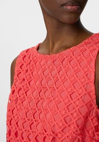 Korallefarbenes ärmelloses Oberteil mit einem strukturierten Rautenmuster, das einen runden Ausschnitt und blumige Stickakzente aufweist. Glatte Stoffoberfläche.