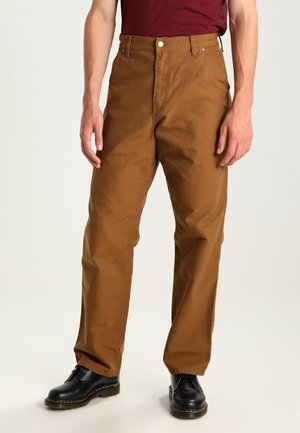 Uomo che indossa pantaloni marroni a gamba dritta, scarpe nere allacciate e una camicia rosso scuro, in piedi davanti a uno sfondo chiaro e uniforme.