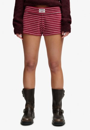 Person iført vinrøde og hvide stribede shorts, mørk vinrød langærmet sweater og mørkebrune midt-på-læggen støvler står med benene let adskilt.
