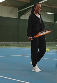 Črni zip-up kombinezon, bele športne čevlje, drži oranžno teniško loparico, na modrem notranjem teniškem igrišču z zelenimi stenami.