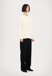 Pull en maille côtelée crème à manches longues, associé à un pantalon noir à larges jambes et des chaussures noires, se tenant devant un fond uni.