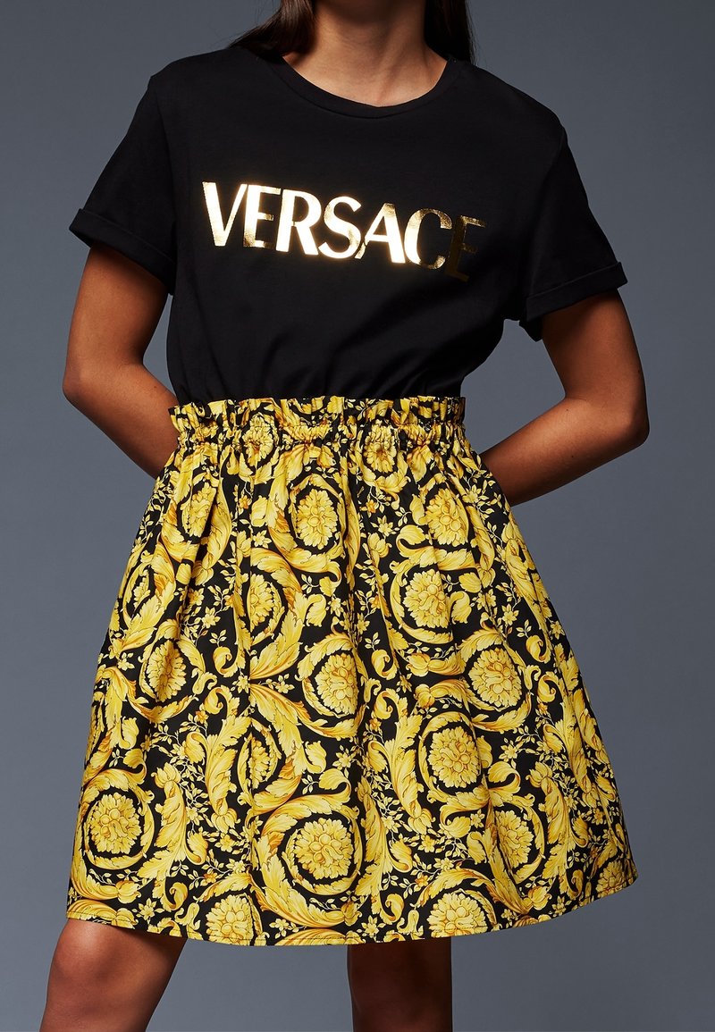 Sort T-shirt med gyldent "VERSACE" tekst kombineret med en højtaljet nederdel med gul og sort udsmykket blomsterbarokmønster.