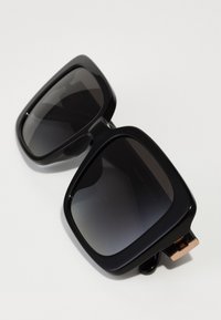 Lunettes de soleil noires surdimensionnées avec des verres teintés sombres, des montures en plastique brillant et un accent doré sur les branches.