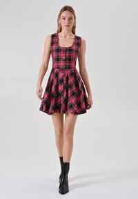 Abito a quadri rosso e nero con un corpetto aderente, scollo quadrato e una gonna ampia e plissettata. Presenta tre bottoni lungo la parte anteriore.