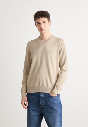 Beige svetr s výstřihem do V vyrobený z měkkého pleteného materiálu, s dlouhými rukávy a žebrovanými manžetami. Model doplněn modrými džíny.