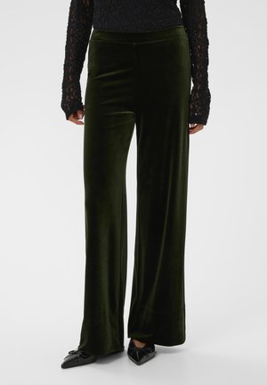 Black lace long-sleeve top paired with high-waisted, wide-leg dark green velvet pants and black pointed-toe flats with bows.